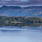 A calm body of water near lush, green foothills with a mountain range in the distance 