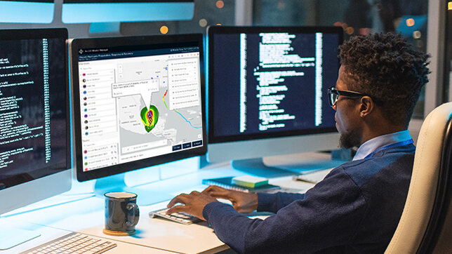 Person sitting at a desk viewing a 2D gray colored map and text on a computer monitor next to two large computer monitors