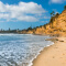 A sunny oceanside beach with foamy blue water, lined with tall cliffs topped with homes and palm trees