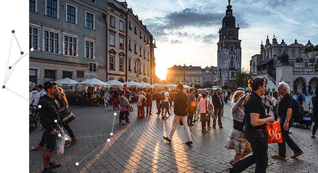 A European square filled with a crowd of people walking and a sidewalk café with outside tables and umbrellas