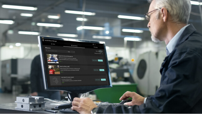 Man with glasses and a blue coat at a desk reviewing a list of ArcGIS Mission operations on a computer with photos and text