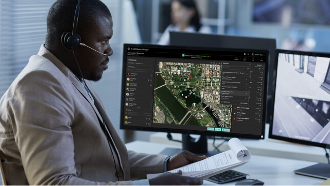 Man at a desk with a headset looking at two computer monitors that show a 2D digital map and a live video feed