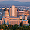 An aerial view of a sprawling city with a mix of modern and historic buildings and rolling blue mountains in the background, warmly lit by a clear sunrise sky