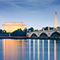 A stately stone bridge at sunset with arches over still blue water with the Lincoln Memorial and Washington Monument lit golden against the darkening blue sky