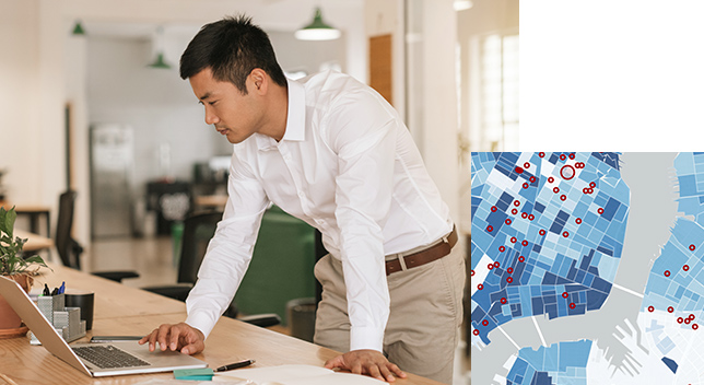 A person wearing business attire leaning on a desk in an open concept office using a laptop overlaid with a graphic map