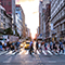 A busy city street with many pedestrians using the crosswalk with upscale storefronts on both sides warmly lit by a golden sunset