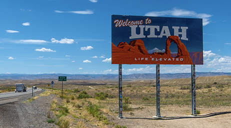 A stretch of interstate highway through wide scrub-covered desert with a large colorful billboard stating “Welcome to Utah” under a bright blue sky