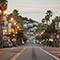 A ground-level view of a San Francisco street lined with shops and palm trees, with terraces of homes and apartment buildings climbing a hillside in the background