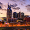 A modern city with high rise buildings brightly lit against a pink and gold sunset sky scattered with dark purple clouds