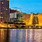 A modern city lit up and reflecting the golden light of sunset beside a bay of sparkling calm water under a clear indigo sky