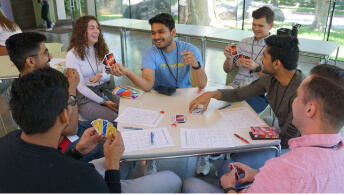 A group of casually dressed people playing a card game around a table in a sunlit modern cafe