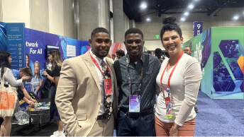 Three professionally-dressed smiling people wearing red conference lanyards and posing in a colorful modern convention center