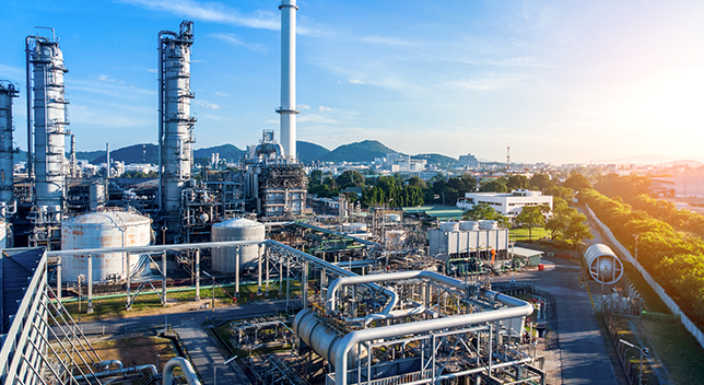 A modern refinery beside a sunrise-lit city and lined with green trees