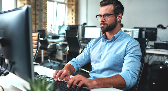 A person in glasses and a light blue collared shirt sitting at a computer workstation in a modern sunlit group office 