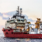 A yellow oil refinery beside choppy blue ocean waters with a red and white oil rig floating alongside beneath a pale overcast sky