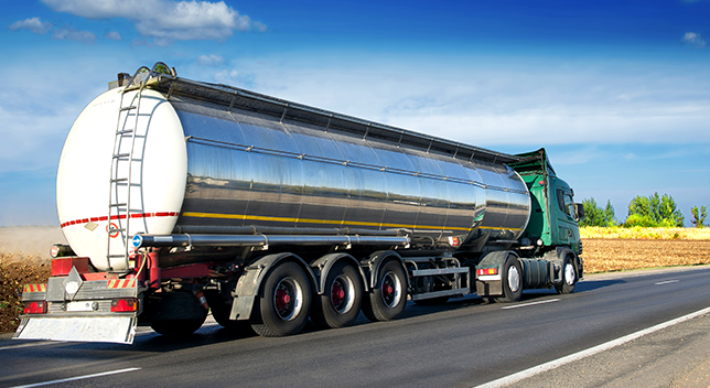 A green and silver big rig driving down a highway beside a large stretch of brown farmland beneath a deep blue sky streaked with clouds