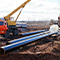 Long shining black pipes nestled in deep freshly-dug dirt trenches alongside utility trucks with cranes under a pale overcast sky