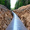 A large gray pipe stretches into the distance, lying inside a rocky trench lined with forest trees under a pale overcast sky