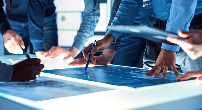 A glowing light table spread with blue maps and surrounded by people wearing long-sleeved buttoned shirts