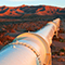 A desert landscape dotted with green scrub and a large white utility pipe stretching into the distance toward orange-lit mountains at sunrise