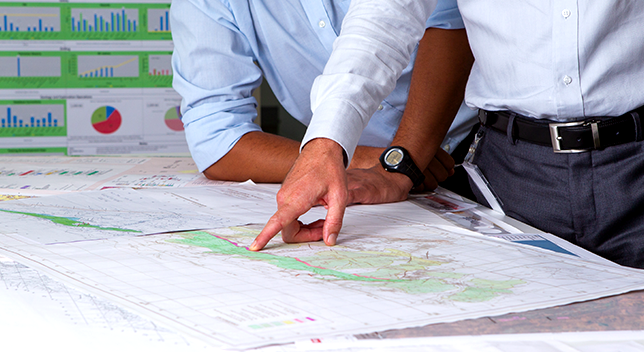 A photo of two professionally-dressed people discussing a table spread with paper maps and charts