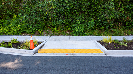 A yellow ADA-compliant curb ramp leading to a sidewalk lined with foliage