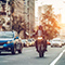 A tree-lined city street lit by a golden sunrise with cars and a motorcyclist in motion in the foreground