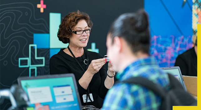 A photo of a conference staffer helping a registrant while a person wearing a backpack works on a laptop in the foreground 