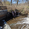 Two workers in waterproof gear handling water measurement equipment beside an overflowing pond