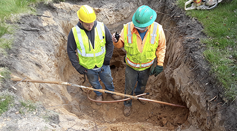 Two mobile workers wearing hard hats and neon yellow vests standing in a shallow ditch