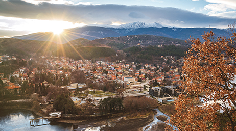 An aerial view of Sofia, Bulgaria with a river, densely populated architecture, and mountains with the sun shining through clouds