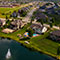 An aerial image of a neighborhood of large houses surrounded by well-kept green lawns beside a large still artificial lake with several fountains