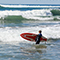 A child standing in the surf on a sunny beach wearing a blue wetsuit and carrying a red surfboard while watching incoming waves