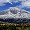 A panoramic view of a sprawling tree-filled suburb with a towering snow-capped mountain range in the background beneath a deep blue sky