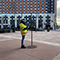 A surveyor wearing a yellow safety vest stands in a paved city square under a hazy sunset sky looking at a clipboard