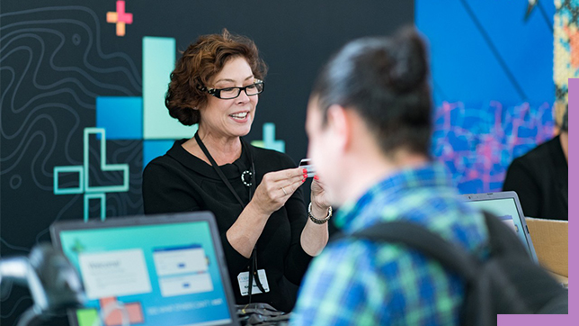 A photo of a conference staffer helping a registrant, while a person in a backpack stands in the foreground using a laptop