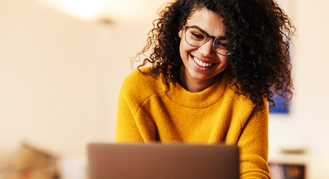 A young person wearing glasses and a yellow sweater smiling as they look at a laptop monitor