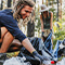 A group of smiling volunteers sitting and crouching on a sun-dappled forest floor gathering trash into large black bags