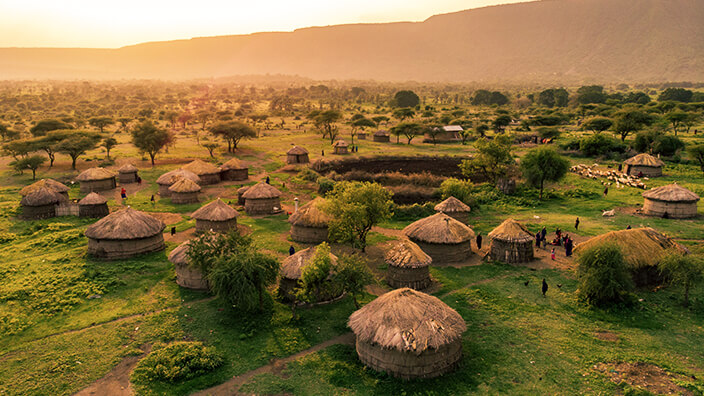 An aerial photo of a village of small brown huts on a green plain with distant mountains silhouetted against a pale orange sunset