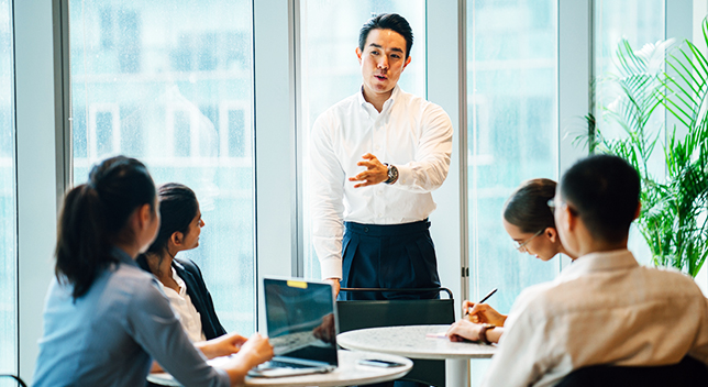 A business person standing in a room speaking with colleagues who are sitting at tables taking notes on laptops and paper