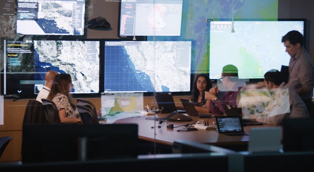 A meeting room filled with people sitting at a table working on laptops and large television screens on the wall displaying maps