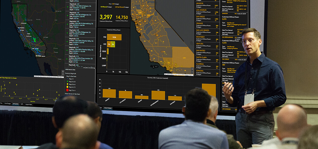 A presenter showing a large screen displaying a dashboard of California to a room full of people sitting down