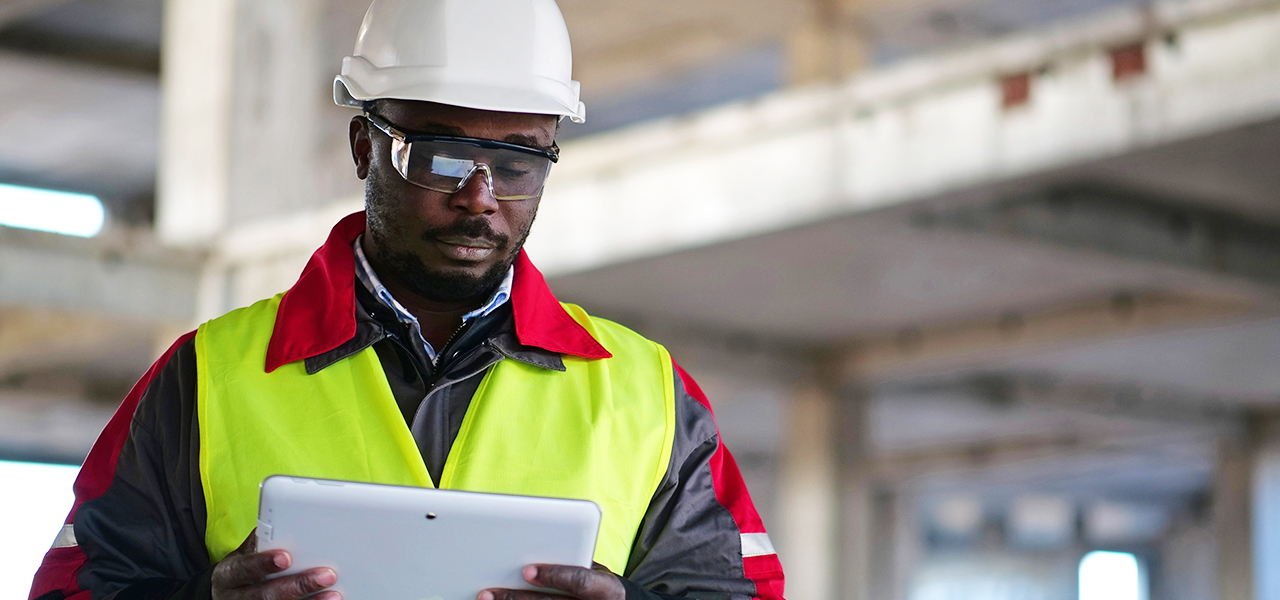 A person wearing a hard hat, safety goggles, and a yellow safety vest using a tablet